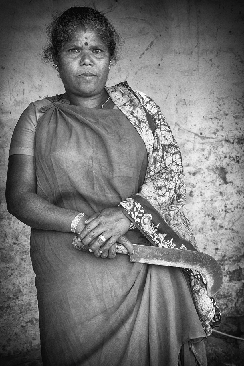 Black and white portrait of a woman in traditional attire holding a sickle, standing confidently against a textured wall — an evocative cultural moment captured by Cristian Mitrani.