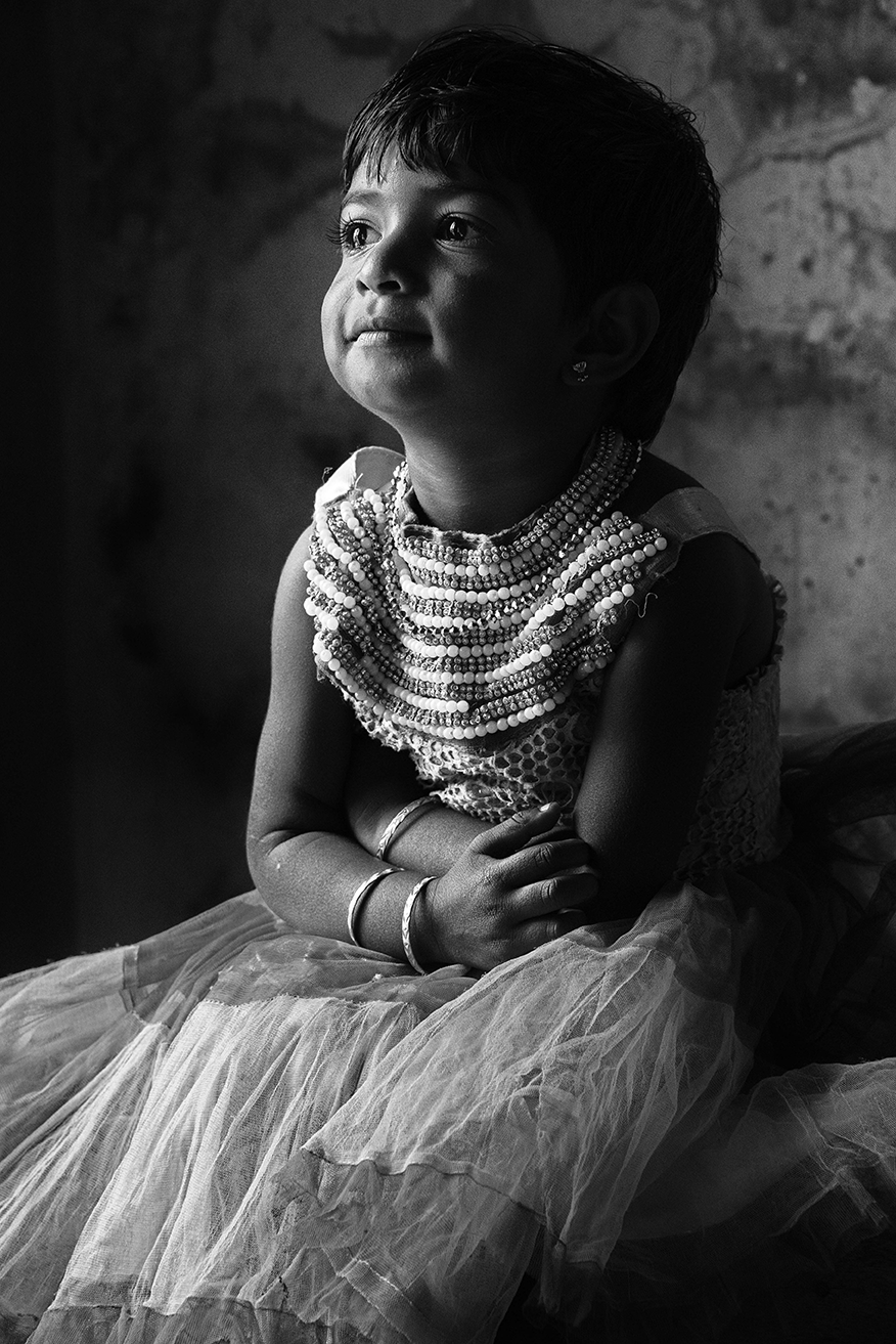 Black and white portrait of a young girl dressed in a tulle skirt and pearl necklace, sitting thoughtfully in soft window light — a delicate and emotive moment captured by Cristian Mitrani.