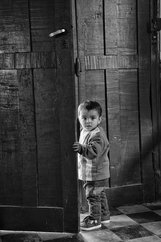 Black and white photo of a small child standing in a doorway, gazing curiously at the camera — a powerful and intimate moment by Cristian Mitrani.