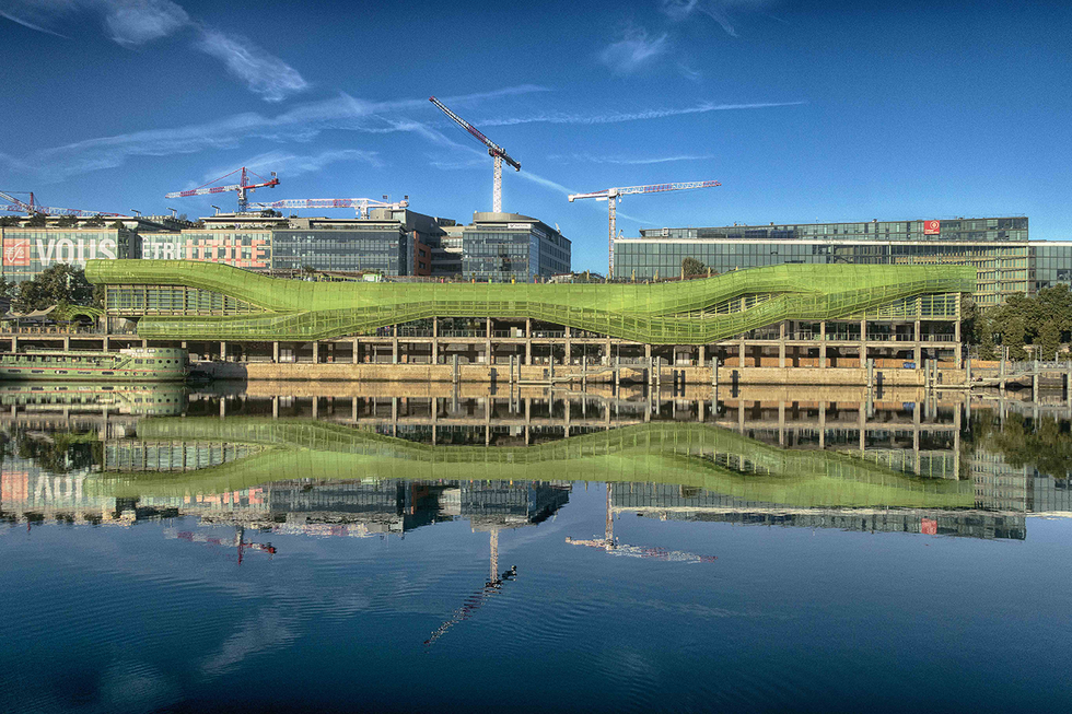 A photograph by Cristian Mitrani capturing a modern construction site along a river, with multiple cranes working atop green-roofed buildings under a bright blue sky with scattered clouds. The scene is reflected in the calm water below, creating a symmetrical mirror image of the construction activity and architecture.