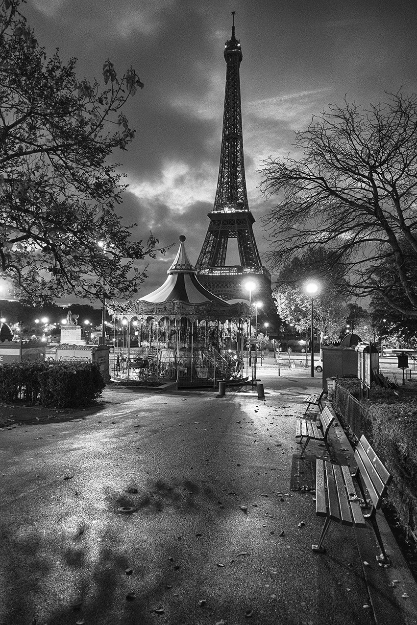 Black-and-white photograph by Cristian Mitrani capturing the Eiffel Tower in Paris at night, with a lively carousel in the foreground and leafless trees framing the scene. The image highlights the illuminated tower against a dark, cloudy sky, creating a nostalgic and timeless atmosphere.