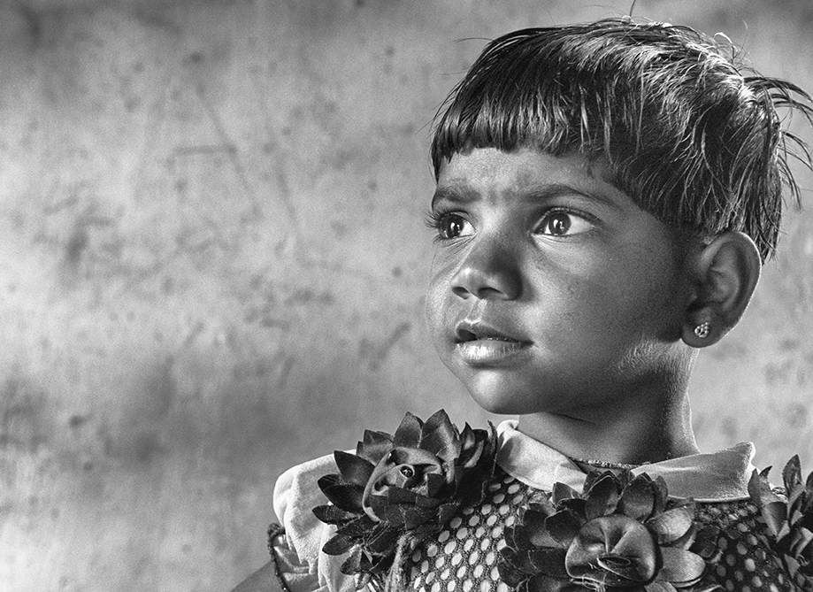 Black and white close-up portrait of a young child looking thoughtfully to the side, adorned with floral decorations — a tender and evocative capture by Cristian Mitrani.