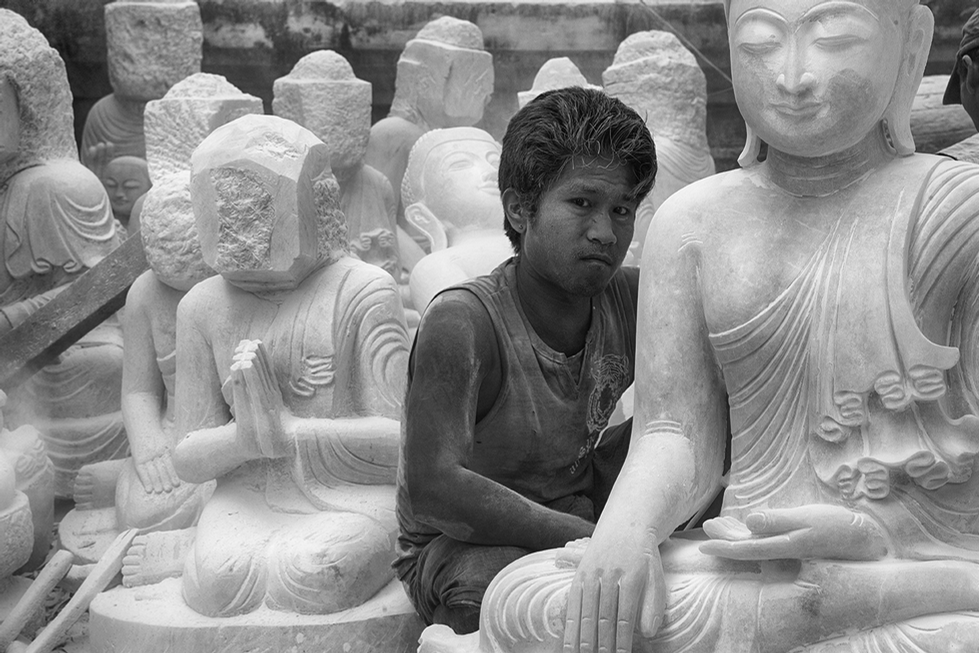 Black and white photo of a young boy sitting among unfinished stone Buddha statues, highlighting contrast between youth and tradition — a thoughtful cultural capture by Cristian Mitrani.
