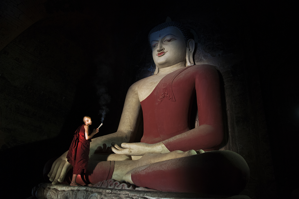 Young monk standing beside a large Buddha statue inside a dimly lit temple, highlighting spiritual devotion — an intimate portrait by Cristian Mitrani.