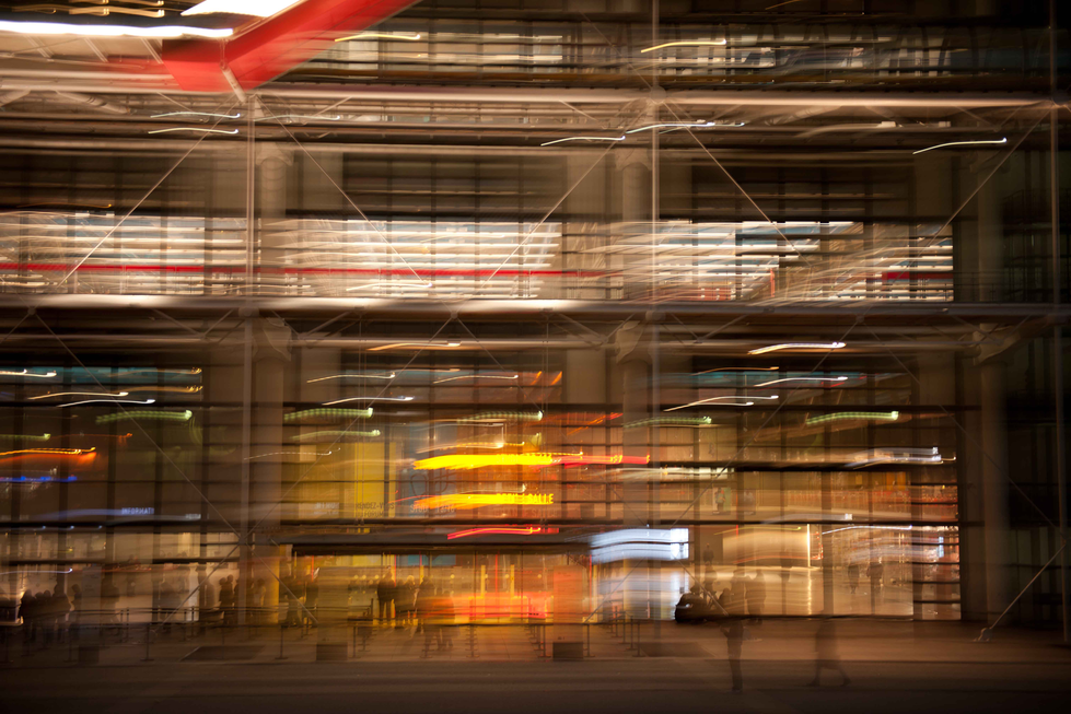 Photograph by Cristian Mitrani capturing a blurred, motion-filled scene of a warehouse or storage facility at night. The image features shelves filled with boxes and goods, with streaks of light suggesting movement and activity within the space, creating an abstract and dynamic representation of industrial or commercial storage.