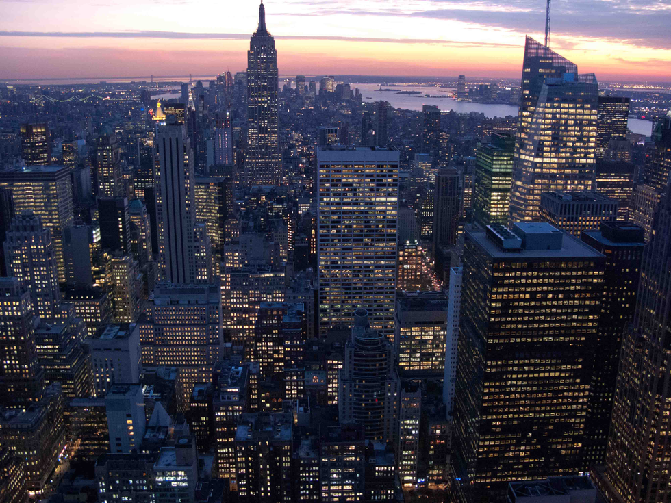 Photograph by Cristian Mitrani capturing a breathtaking aerial view of New York City skyline at dusk. The image features illuminated skyscrapers, including the Empire State Building, with the city lights reflecting off the windows. In the background, the sky transitions from sunset colors to night, with the distant horizon and water visible, showcasing the vibrant urban landscape.
