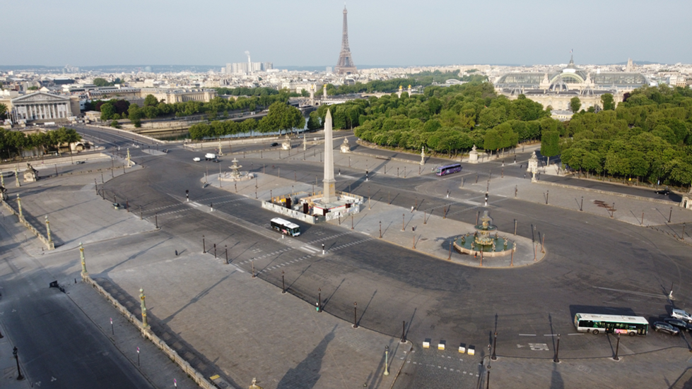 A photograph by Cristian Mitrani capturing a panoramic view of Place Charles de Gaulle in Paris, featuring the iconic Arc de Triomphe at the center. The wide circular road is lined with trees and ornate street lamps, with a few vehicles visible on the streets. The Eiffel Tower stands prominently in the background under a clear sky, showcasing Mitrani's signature dramatic and textured photographic style.