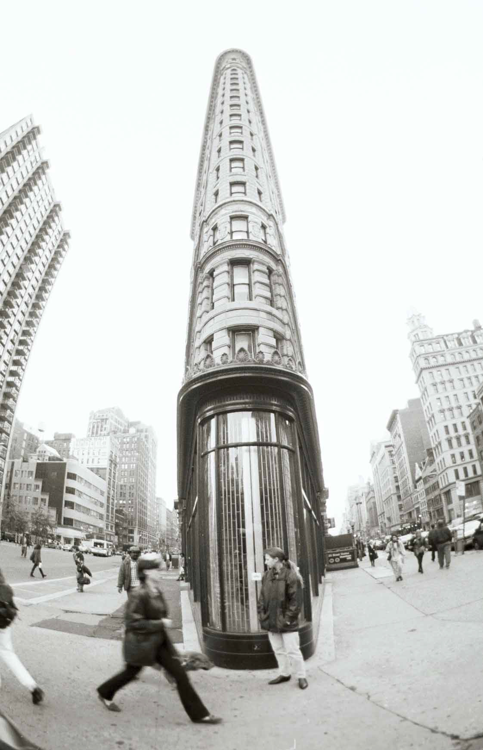 Photograph by Cristian Mitrani depicting the iconic Flatiron Building in New York City. The image is captured from a low angle, showcasing the tall, triangular-shaped skyscraper surrounded by busy streets with pedestrians and vehicles, highlighting the urban atmosphere and architectural prominence of the structure.