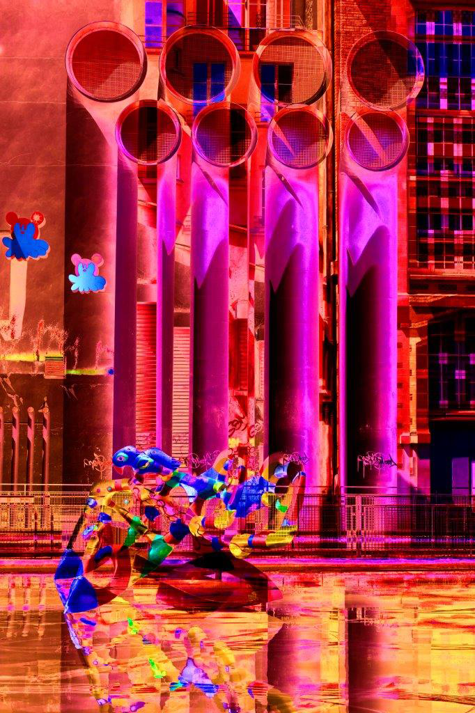 An abstract, vibrant photograph by Cristian Mitrani featuring a series of tall, glowing columns with circular openings, illuminated in shades of pink, purple, and red. The scene includes reflections on a glossy surface below, and geometric patterns in the background, creating a dynamic and surreal composition characteristic of Mitrani's experimental and colorful style.
