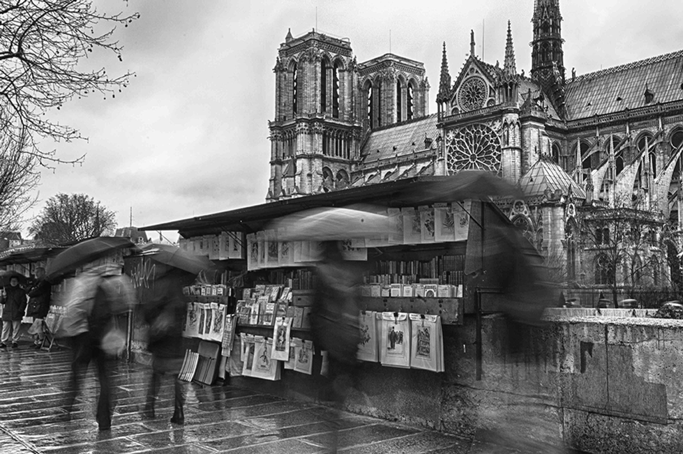 Black-and-white photograph by Cristian Mitrani capturing a street scene featuring a historic Gothic cathedral in the background, with intricate architectural details and tall spires. In the foreground, a street vendor's kiosk displays various posters and newspapers, with blurred pedestrians passing by, emphasizing the lively urban atmosphere and the contrast between the old architecture and everyday city life.