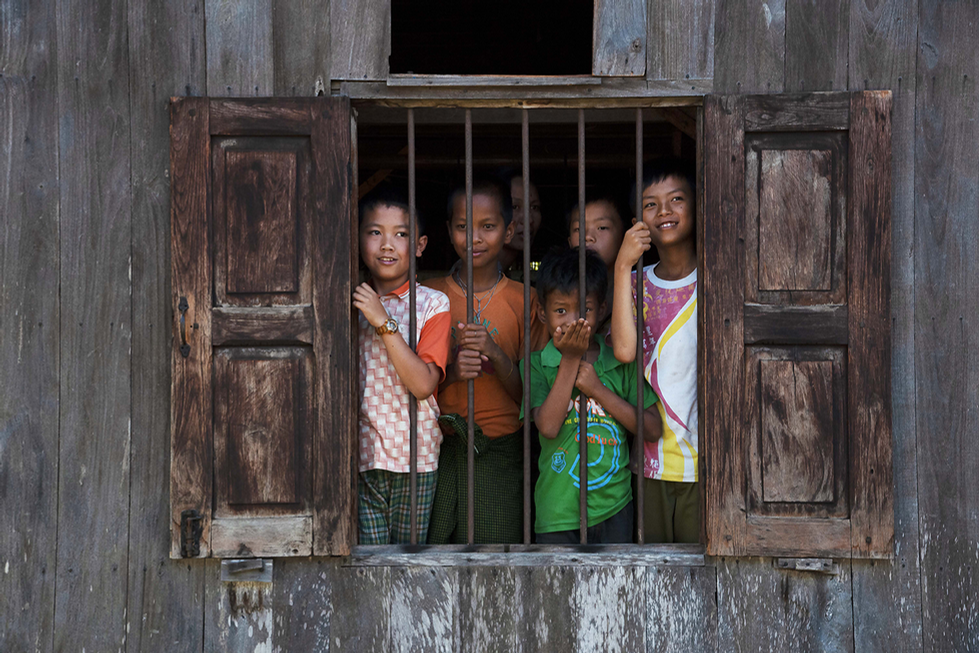Group of children looking through barred wooden window of a rustic building, their expressions curious and hopeful — a poignant social scene captured by Cristian Mitrani.
