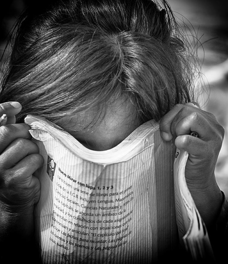 Black and white close-up of a child hiding their face behind a piece of printed paper, hands gripping the edges — a poignant and intimate moment captured by Cristian Mitrani.