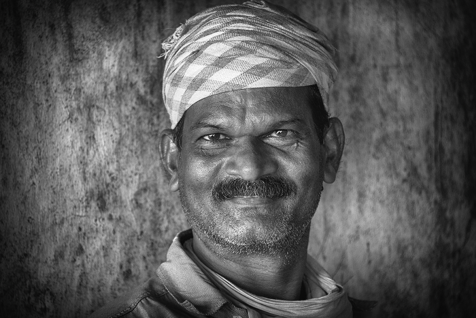 Black and white portrait of a smiling man wearing a checkered turban and scarf, standing against a textured wall — a warm and candid capture by Cristian Mitrani.