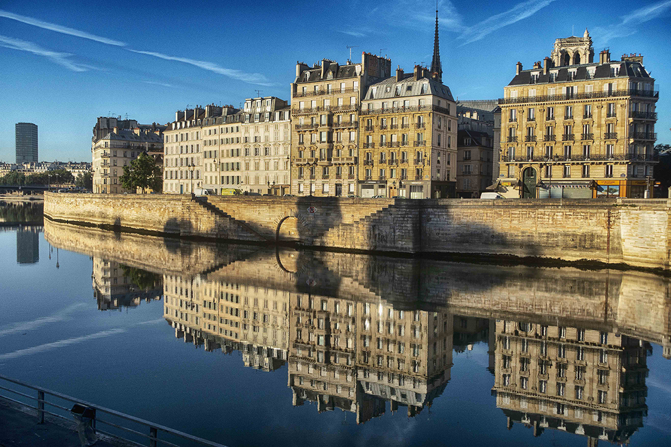 A photograph by Cristian Mitrani showcasing a row of historic Parisian buildings along the Seine River, with their reflections perfectly mirrored in the calm water. The buildings feature classic Parisian architecture with mansard roofs and ornate facades, under a bright blue sky with some clouds.