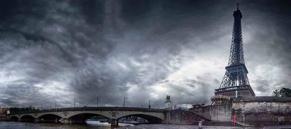 A black-and-white photograph by Cristian Mitrani capturing the Eiffel Tower in Paris, France, from across the River Seine. The scene is dramatic, with dark, swirling clouds in the sky that create a moody atmosphere. The Eiffel Tower stands prominently on the right side of the image, with the river and bridge in the foreground, exemplifying Mitrani's signature style of atmospheric urban and architectural photography that emphasizes mood and intricate details.