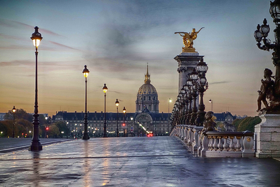 Photograph by Cristian Mitrani of the Alexandre III Bridge in Paris during early evening, featuring the ornate golden statues atop the bridge's pillars. The scene shows the illuminated lamps along the bridge, with the domed Les Invalides building in the background against a moody, cloudy sky, and the wet surface of the bridge reflecting the lights.