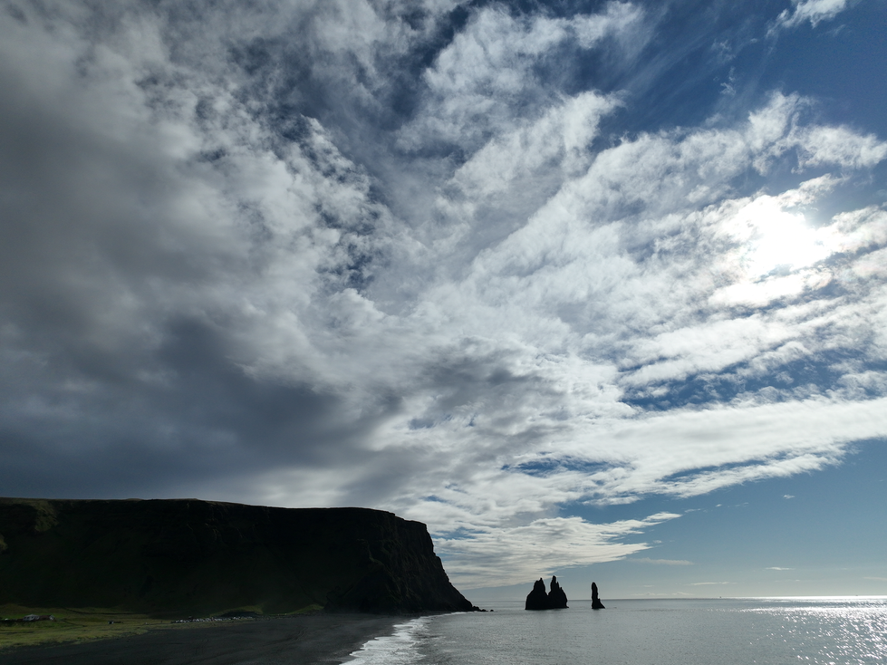 An aerial photograph by Cristian Mitrani capturing a dramatic coastal scene with dark cliffs on the left and three prominent sea stacks rising from the ocean on the right. The sky is filled with dynamic, textured clouds, and the sunlight breaks through, illuminating parts of the scene. Mitrani's signature style emphasizes the contrast and depth in this striking landscape.