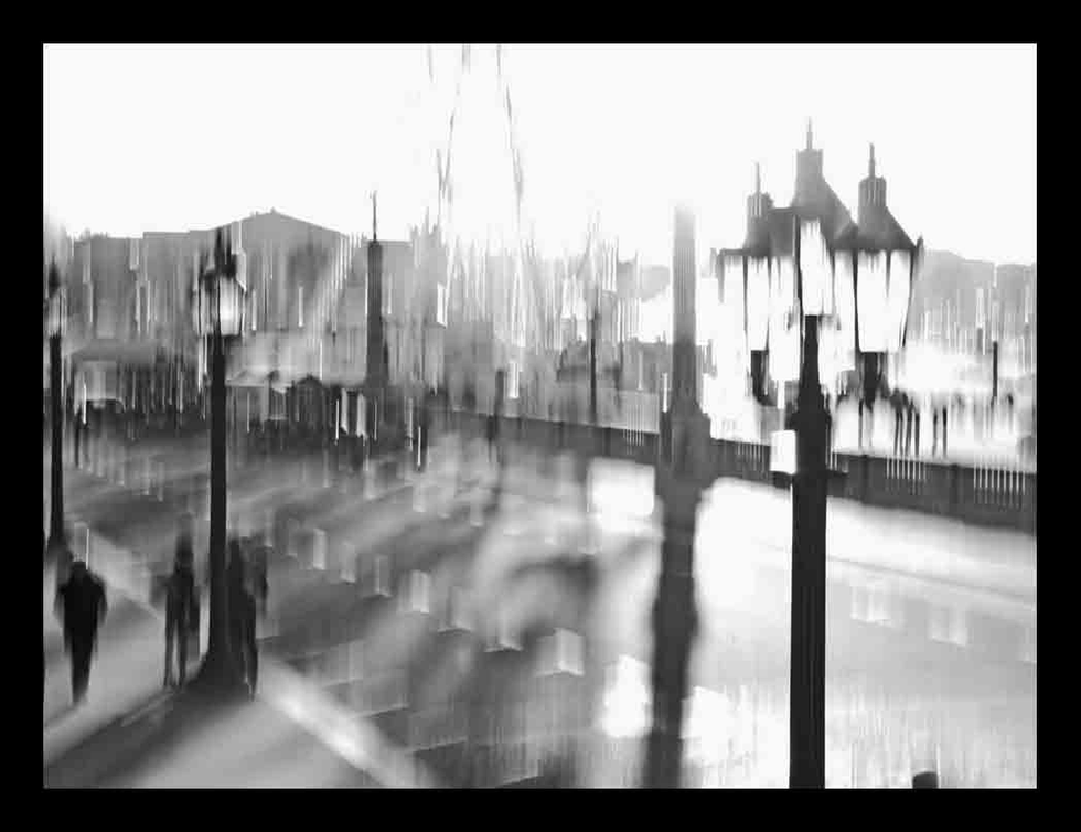 High-contrast black and white photo of a riverside promenade with lamp posts, blurred figures, and radiant sunlight — a poetic urban scene by Cristian Mitrani.