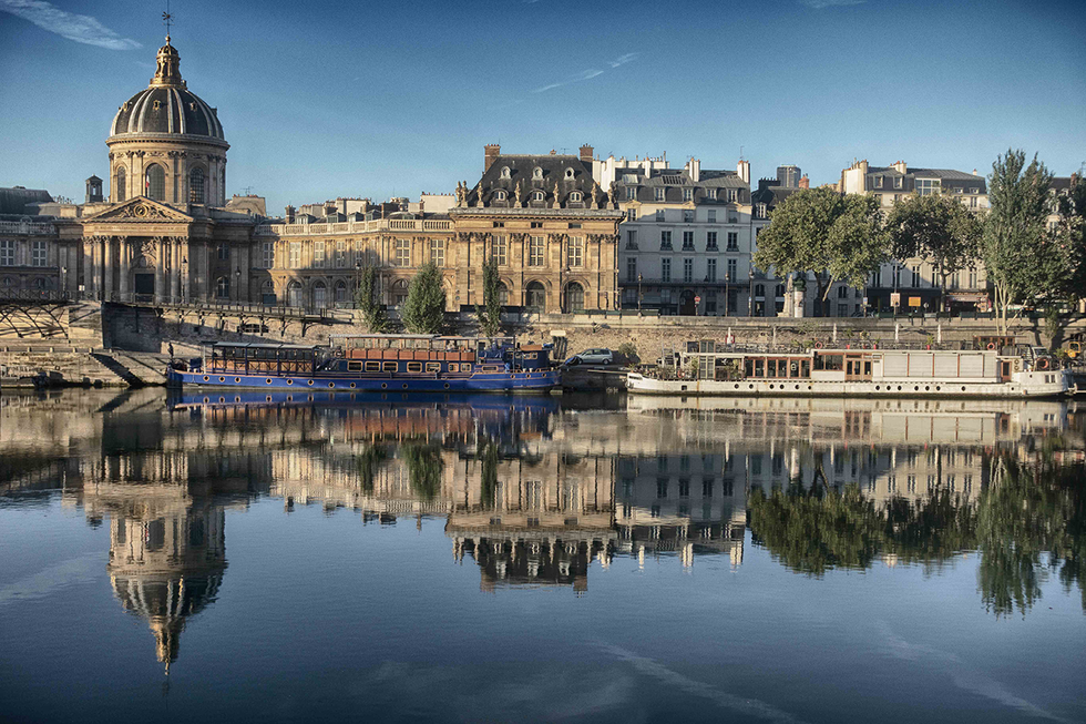 Photograph by Cristian Mitrani capturing a panoramic view of Paris along the Seine River, featuring historic buildings and the Institut de France with its dome on the left. The calm river water creates a mirror-like reflection of the cityscape under a clear blue sky.