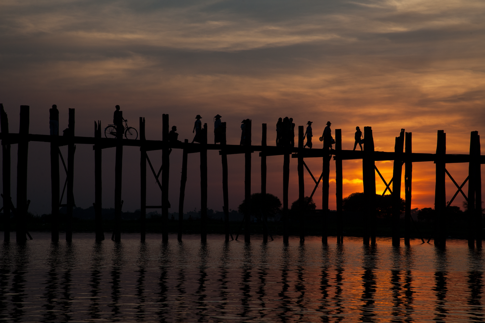 An aerial photograph by Cristian Mitrani capturing a silhouette of people walking across a wooden bridge at sunset. The sky is painted with warm hues of orange and purple, reflected beautifully on the calm water below. The scene emphasizes the tranquil, serene atmosphere and the human connection against the vibrant sky.