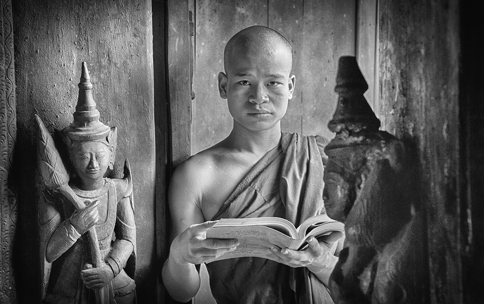 Black and white portrait of a young monk reading a book, surrounded by traditional statues, captured with calm focus — a serene cultural study by Cristian Mitrani.