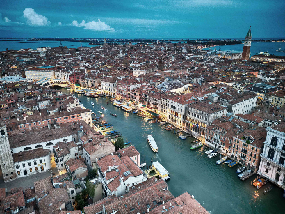 An aerial view of a historic European city with a winding canal lined by colorful buildings and boats, set against a backdrop of a distant horizon and blue sky. This scene highlights Cristian Mitrani's talent for capturing vibrant urban landscapes and the charm of historic architecture.