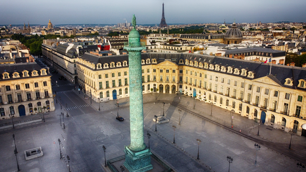 A photograph by Cristian Mitrani capturing a high-angle view of a historic Parisian square featuring the prominent Vendôme Column in the foreground. Surrounding the square are elegant, classical buildings with symmetrical facades. In the background, the Eiffel Tower rises above the city skyline under a clear sky, showcasing Mitrani's characteristic detailed and textured photographic style.