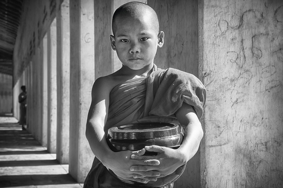 Young Buddhist monk standing in a corridor holding a bowl, bathed in soft light — a serene cultural portrait by Cristian Mitrani.