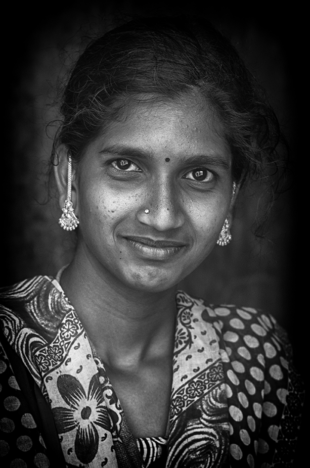 Black and white portrait of a young woman with gentle smile, adorned with traditional earrings and patterned clothing — an intimate cultural moment by Cristian Mitrani.