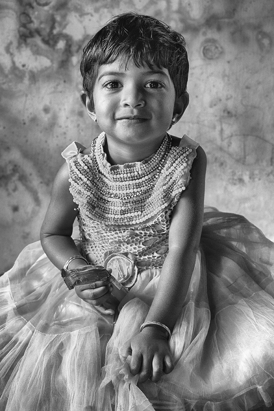 Black and white portrait of a young girl in a tulle dress smiling gently at the camera, sitting against a textured wall — a warm and intimate capture by Cristian Mitrani.