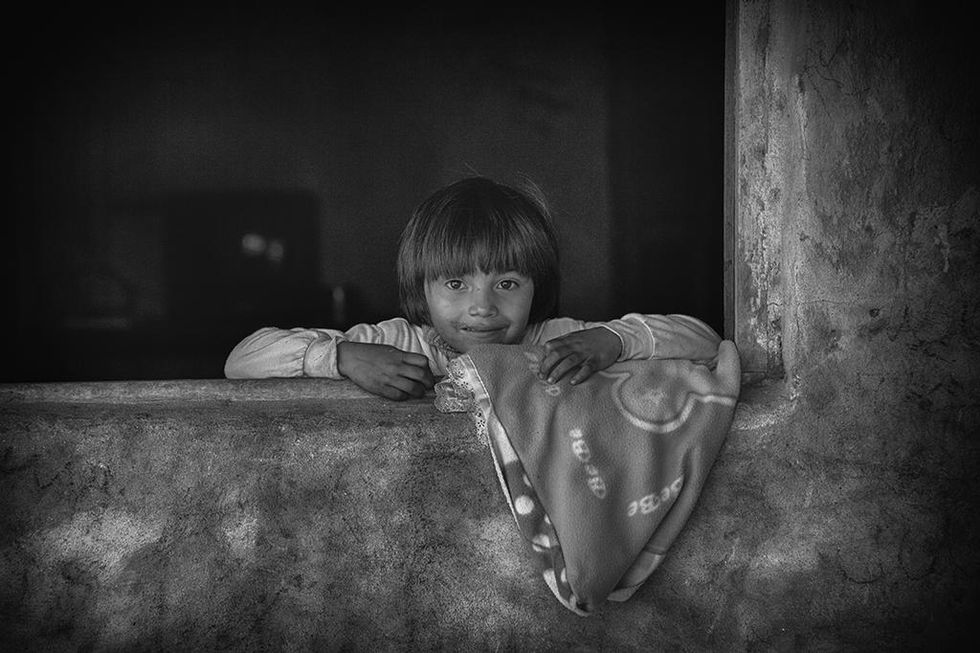 Black and white photo of a child leaning on a windowsill, holding a patterned blanket with a gentle smile — an emotive scene by Cristian Mitrani.