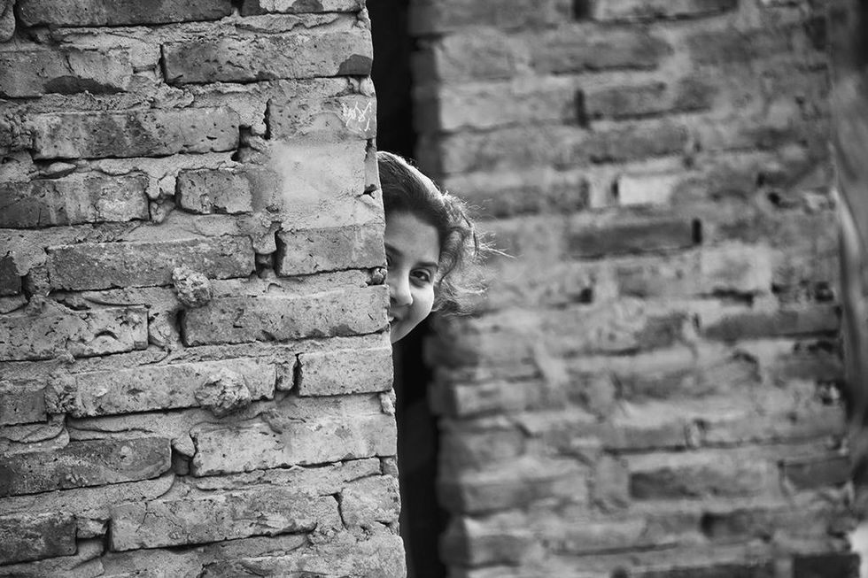 Black and white portrait of a smiling girl peeking from behind a brick wall — a candid and intimate moment captured by Cristian Mitrani.