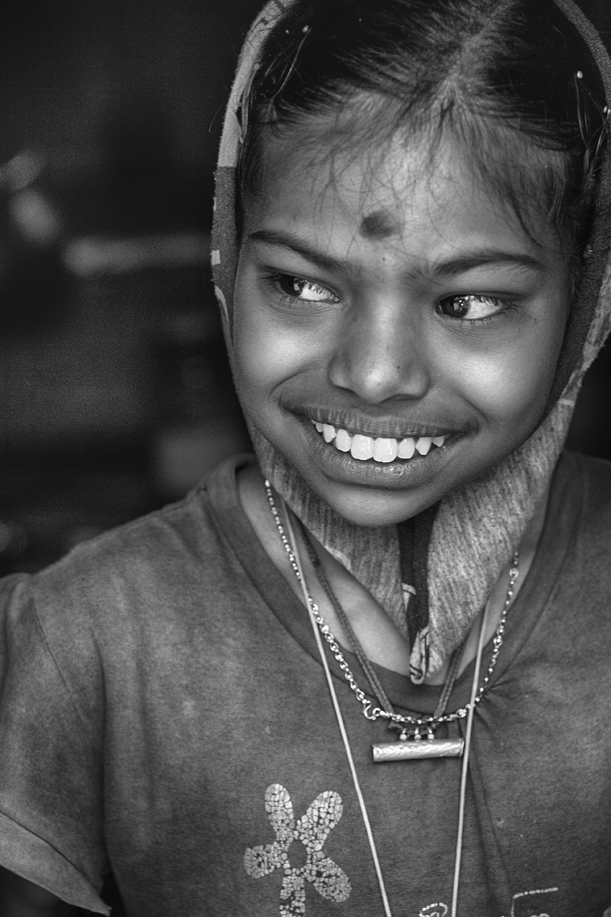 Black and white candid portrait of a smiling young girl wearing a headscarf and layered necklaces, expressing joy and innocence — a heartfelt capture by Cristian Mitrani.