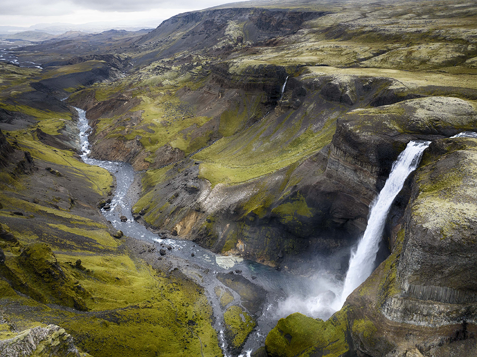 A breathtaking landscape photograph by Cristian Mitrani depicting a rugged canyon with steep cliffs covered in lush green moss. A powerful waterfall cascades down into a winding river below, surrounded by dramatic terrain and atmospheric mist, highlighting the raw beauty of nature.
