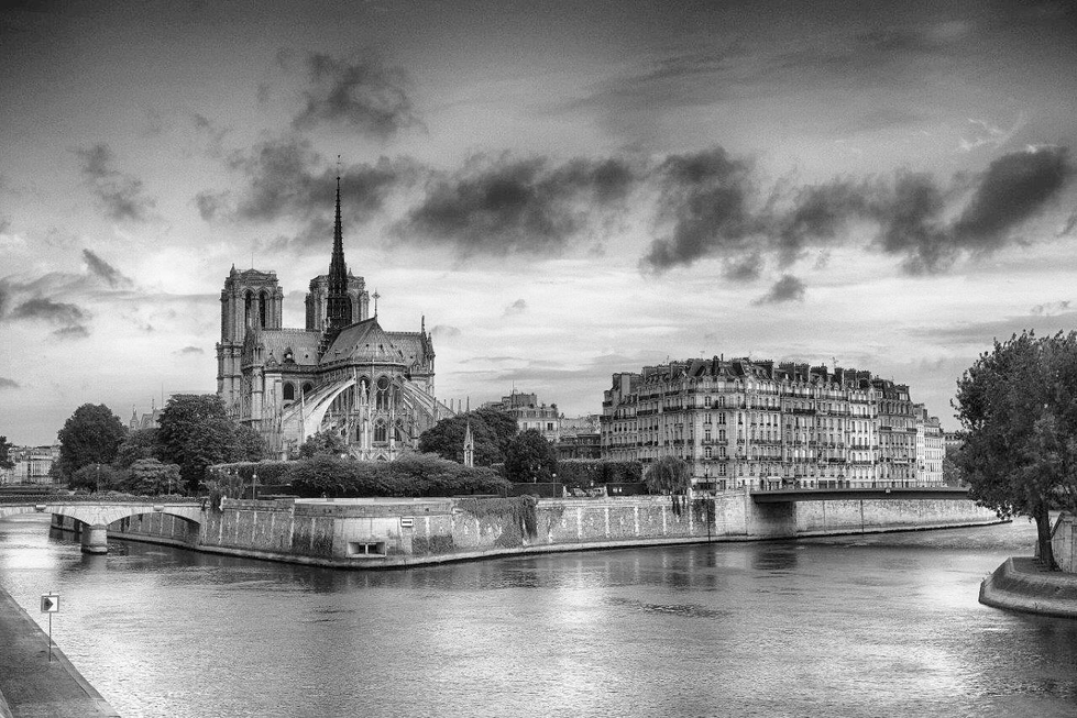 Black-and-white photograph by Cristian Mitrani capturing the Notre-Dame Cathedral in Paris, with its iconic spire and Gothic architecture prominently visible. The Seine River flows in the foreground, reflecting the cloudy sky above. Historic buildings line the riverbank, and the overall scene exudes a timeless, atmospheric quality characteristic of Mitrani's urban photography.