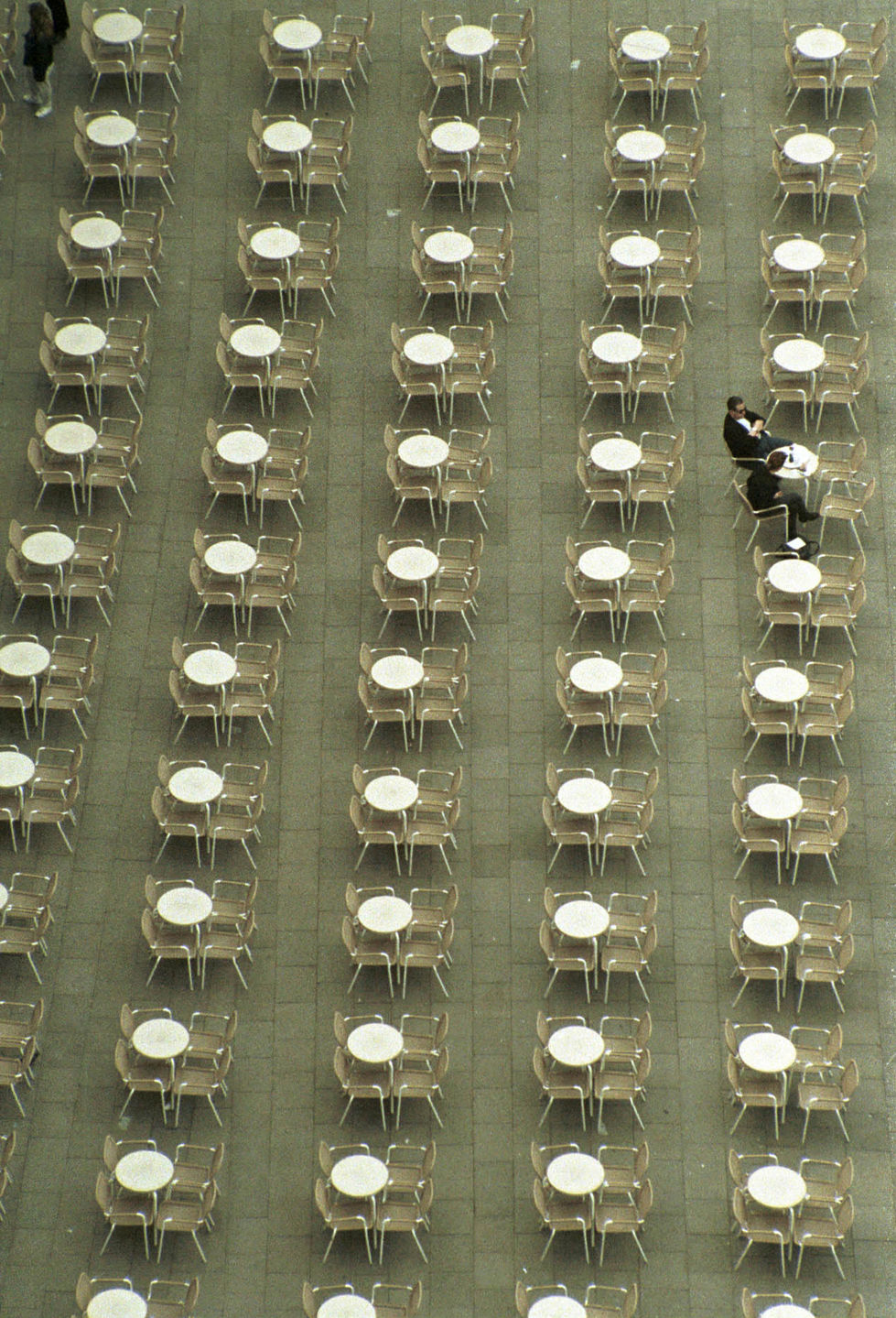 An overhead shot of a large, organized space filled with rows of white chairs arranged in a grid pattern. In the upper right section, a solitary person dressed in black is walking among the empty chairs. The photograph showcases Cristian Mitrani's signature use of symmetry, pattern, and perspective, emphasizing the uniformity and solitude within the space.