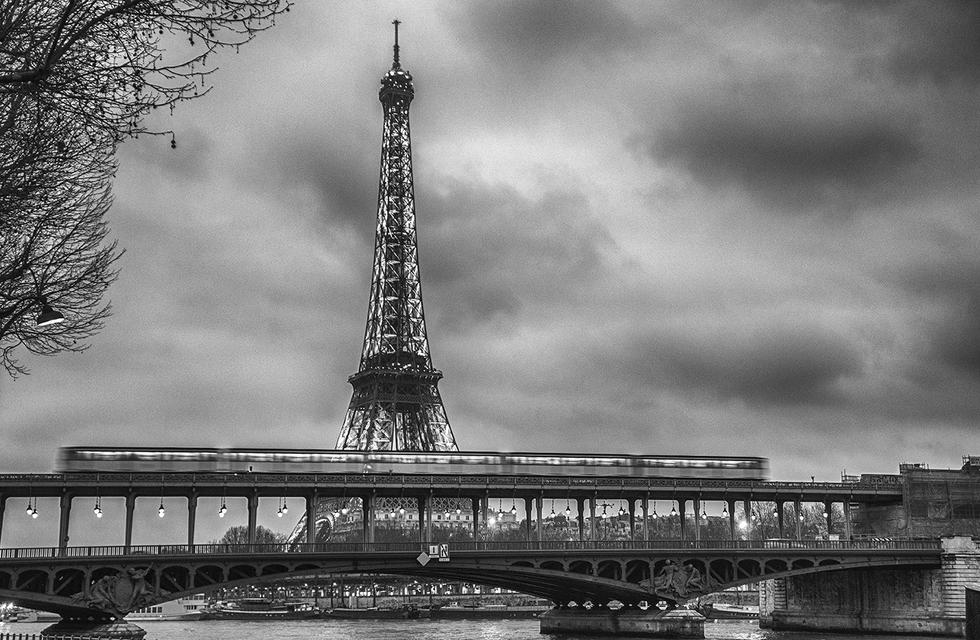Black-and-white photograph by Cristian Mitrani of the Eiffel Tower in Paris, viewed from below the Pont de Bir-Hakeim bridge. The image captures the iconic iron structure towering against a cloudy sky, with the bridge's ornate details and the River Seine visible in the foreground.