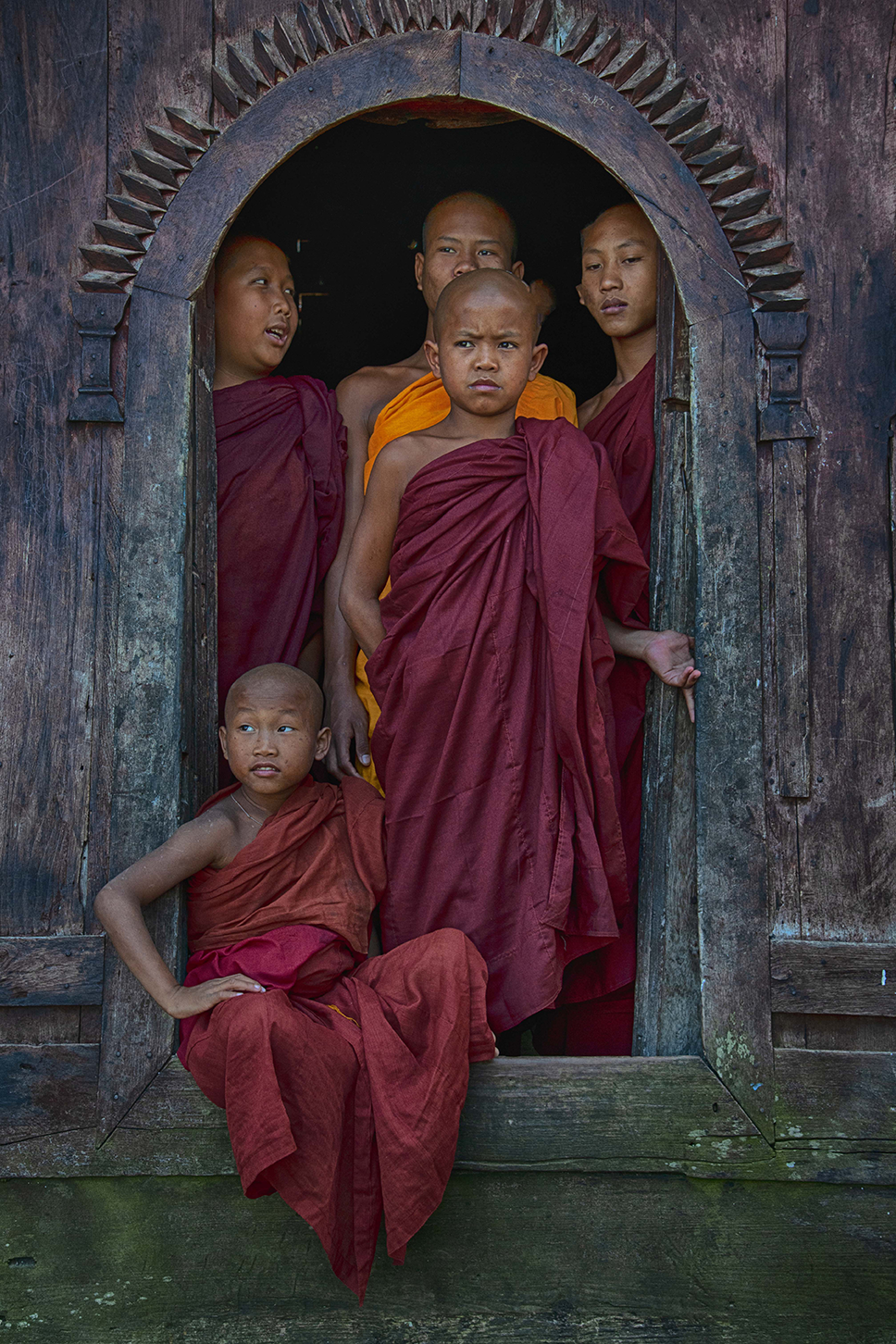 Group of young Buddhist monks in traditional robes standing framed by a carved wooden doorway, displaying calm and introspective expressions — a serene cultural portrait by Cristian Mitrani.