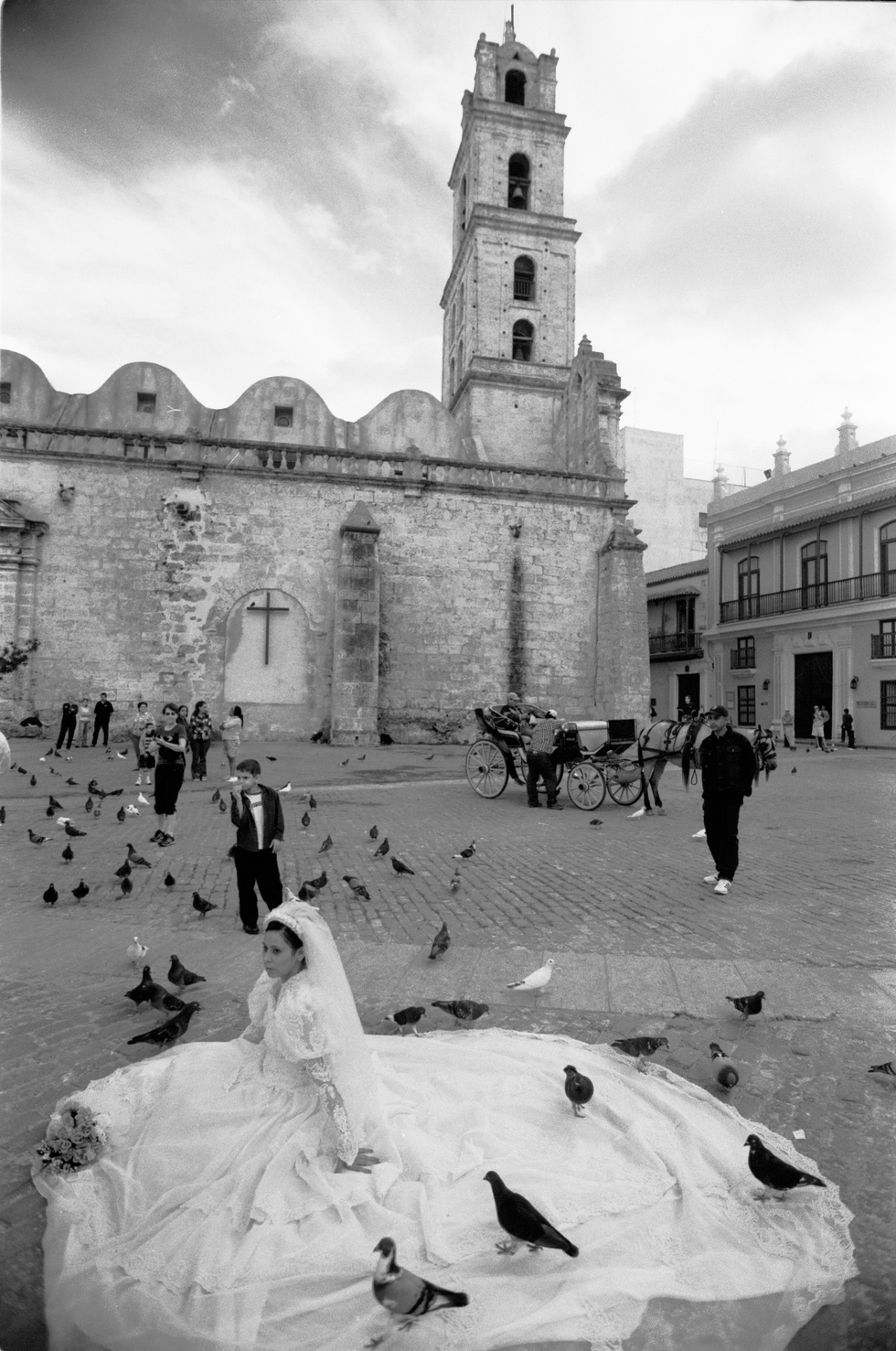 Black-and-white photograph by Cristian Mitrani depicting a lively square with a historic stone church featuring a tall bell tower in the background. In the foreground, a woman in a wedding dress sits on the ground surrounded by pigeons, with some pigeons perched on her dress. People are walking and socializing around the square, capturing a candid and vibrant moment in an urban setting.