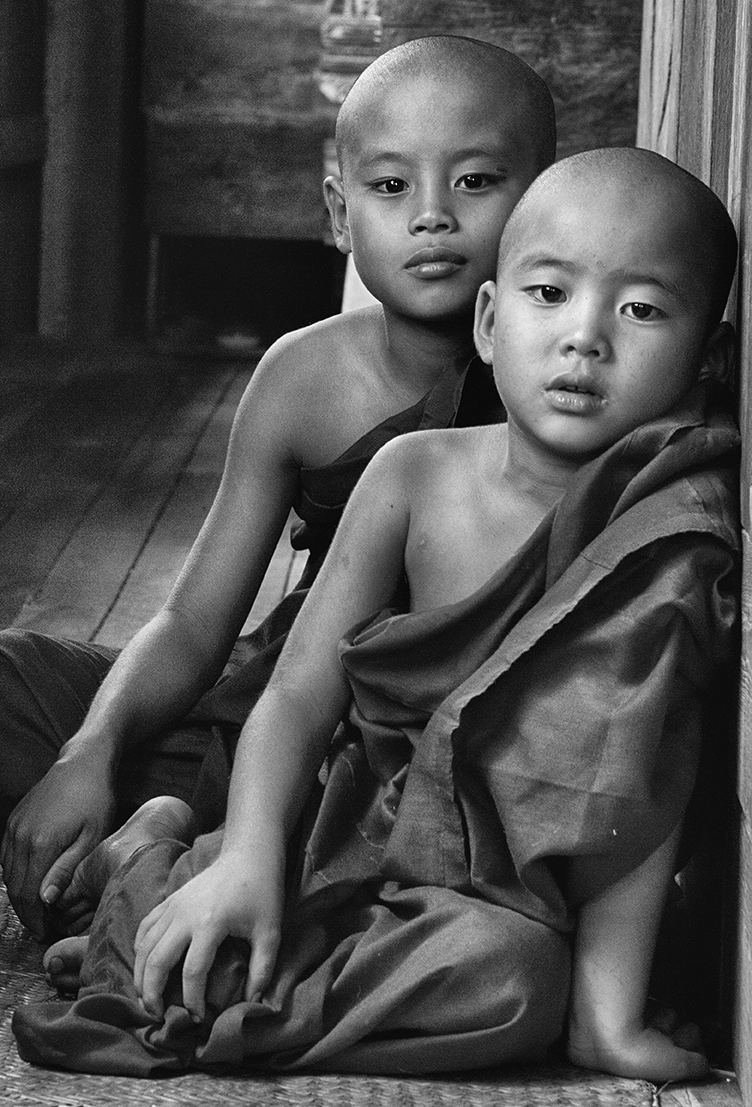 Black and white portrait of two young Buddhist monks sitting closely together, their expressions calm and contemplative — a serene cultural moment captured by Cristian Mitrani.
