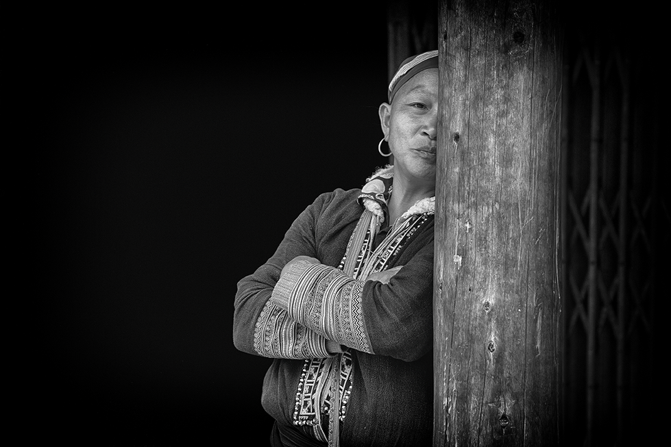 Black and white portrait of a woman standing beside a wooden pillar, her expression calm and reflective — a thoughtful moment captured by Cristian Mitrani.