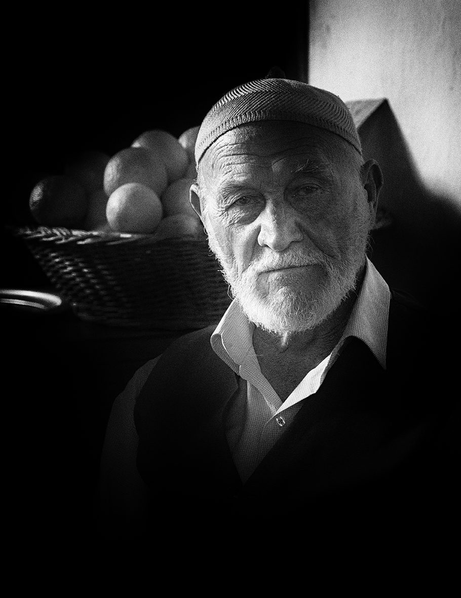 Black and white portrait of an elderly man wearing a cap, softly lit with a basket of fruit in the background — a serene and intimate capture by Cristian Mitrani.
