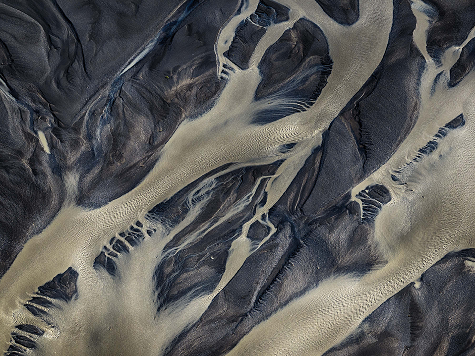 An aerial photograph by Cristian Mitrani depicting a river delta with intricate, winding channels flowing through dark, textured landmasses. The water appears milky or foamy, creating a striking contrast with the surrounding terrain. Mitrani's style emphasizes the detailed patterns and dynamic flow of the waterways, highlighting the natural complexity of the landscape.