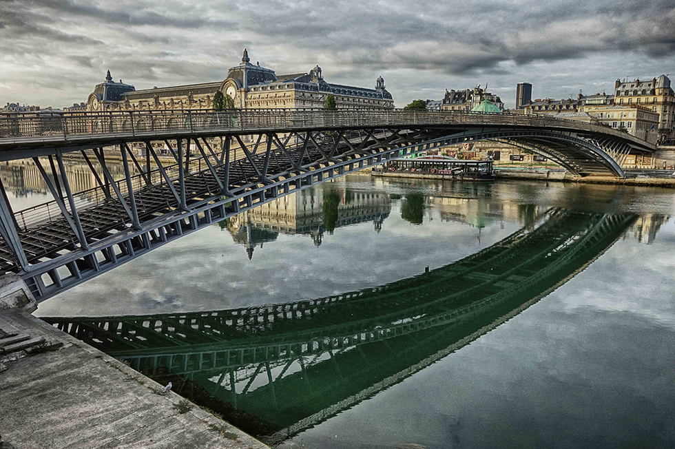 Photograph by Cristian Mitrani capturing the Pont Alexandre III bridge in Paris reflected in the calm waters of the River Seine. The image shows the ornate bridge structure with its decorative lamps and sculptures, set against a cloudy sky with historic Parisian buildings in the background. The reflection creates a symmetrical and serene scene.