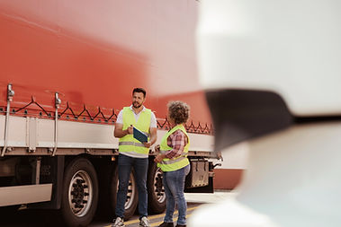Two workers in yellow vests stand by a red truck, discussing a clipboard. They're in a parking lot, with a white vehicle in the foreground.