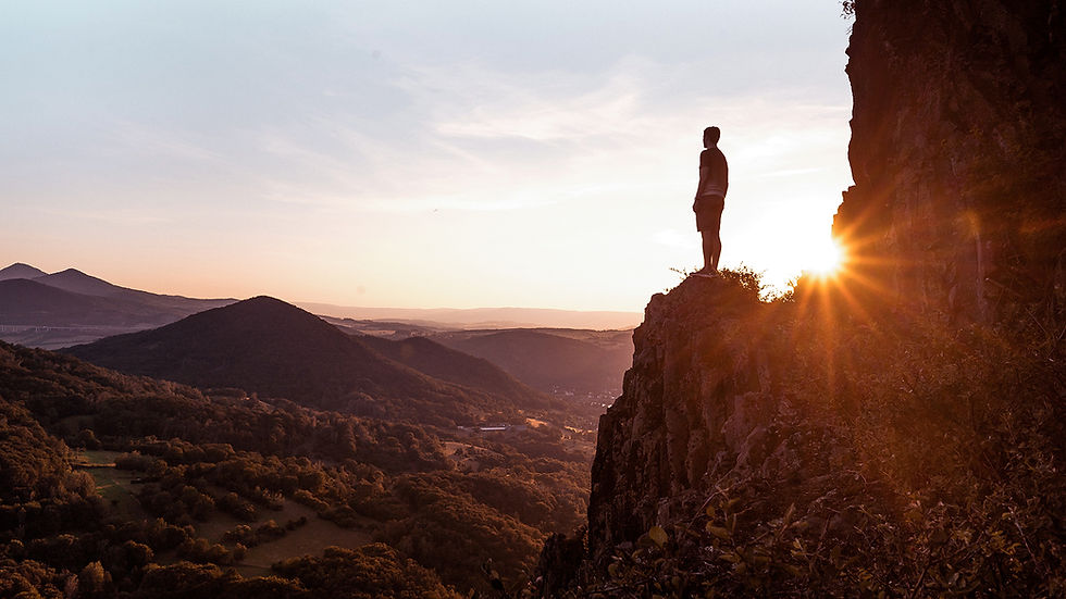 A man standing on a high cliff at sunrise looks out over a forested valley with hills and a city in the distance