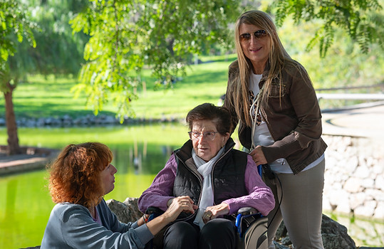 two-women-supporting-elderly-mother-wheelchair-by-lake-park.jpg