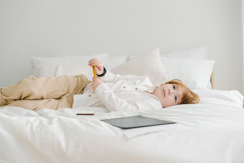 Un bambino autistico con capelli rossi giace rilassato in un letto con lenzuola bianche, tenendo in mano una matita gialla. Accanto a lui sul letto, c'è un cartoncino nero. L'ambientazione è luminosa e tranquilla, suggerendo un momento di riflessione o di pausa creativa.