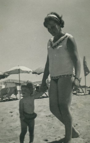 Nella foto in bianco e nero, una donna e un bambino piccolo camminano mano nella mano sulla spiaggia. La donna guarda verso il bambino con un'espressione premurosa, mentre il bambino guarda avanti verso la fotocamera. Entrambi sono a piedi nudi sulla sabbia e sembrano essere in un momento di tranquillità e connessione. Sullo sfondo, si intravedono ombrelloni da spiaggia e sdraio, indicando una giornata tipica al mare.