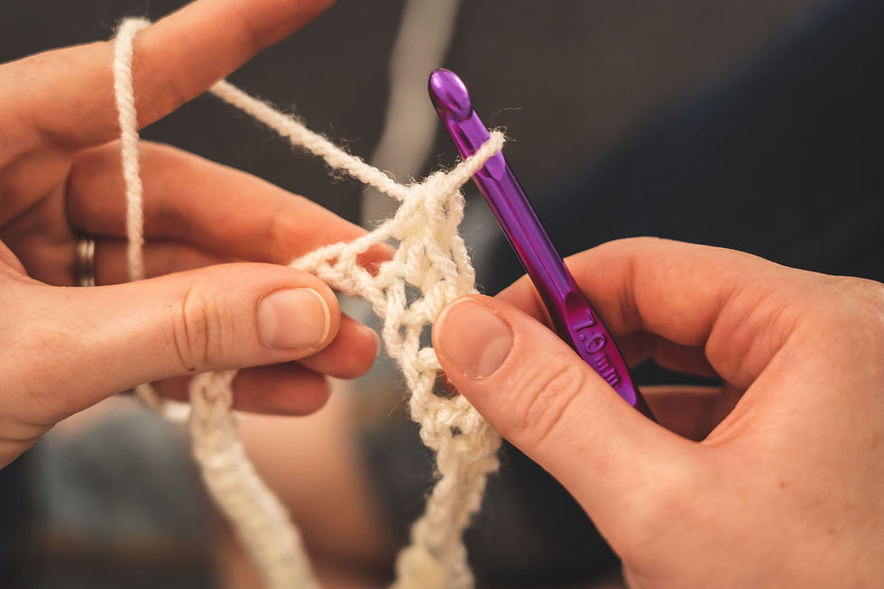 L'immagine mostra le mani di una persona intenta a lavorare a uncinetto. Sta utilizzando un filato bianco e un uncinetto di colore viola. L'uncinetto, specificamente, ha una dimensione indicata su di esso: '10 mm'. Le mani sono in primo piano, mostrando con precisione la tecnica di lavorazione e il filato. La persona ha le unghie curate e indossa un anello, suggerendo un senso di cura e attenzione ai dettagli. Il background è sfocato, permettendo di focalizzare l'attenzione sulle mani e sull'uncinetto. L'immagine trasmette un'atmosfera calma e rilassante, tipica dei momenti dedicati all'artigianato e alla creatività.